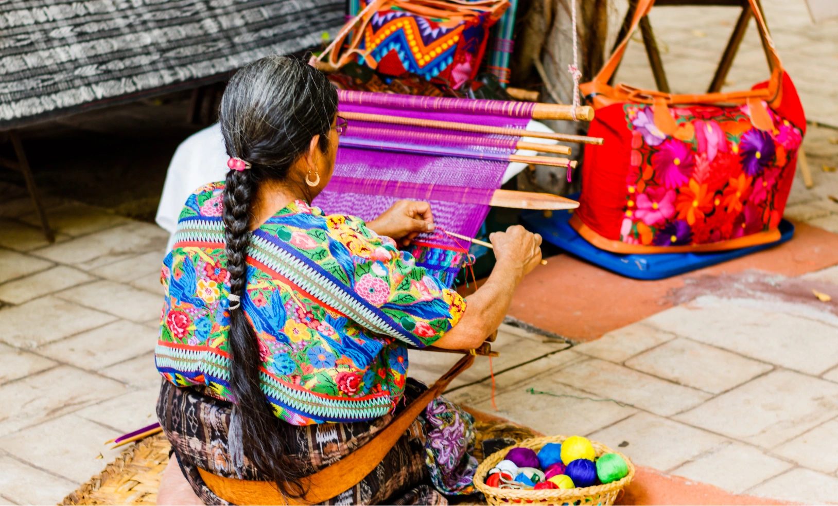 A woman in traditional colorful attire sitting and skillfully weaving a vibrant purple and red textile.