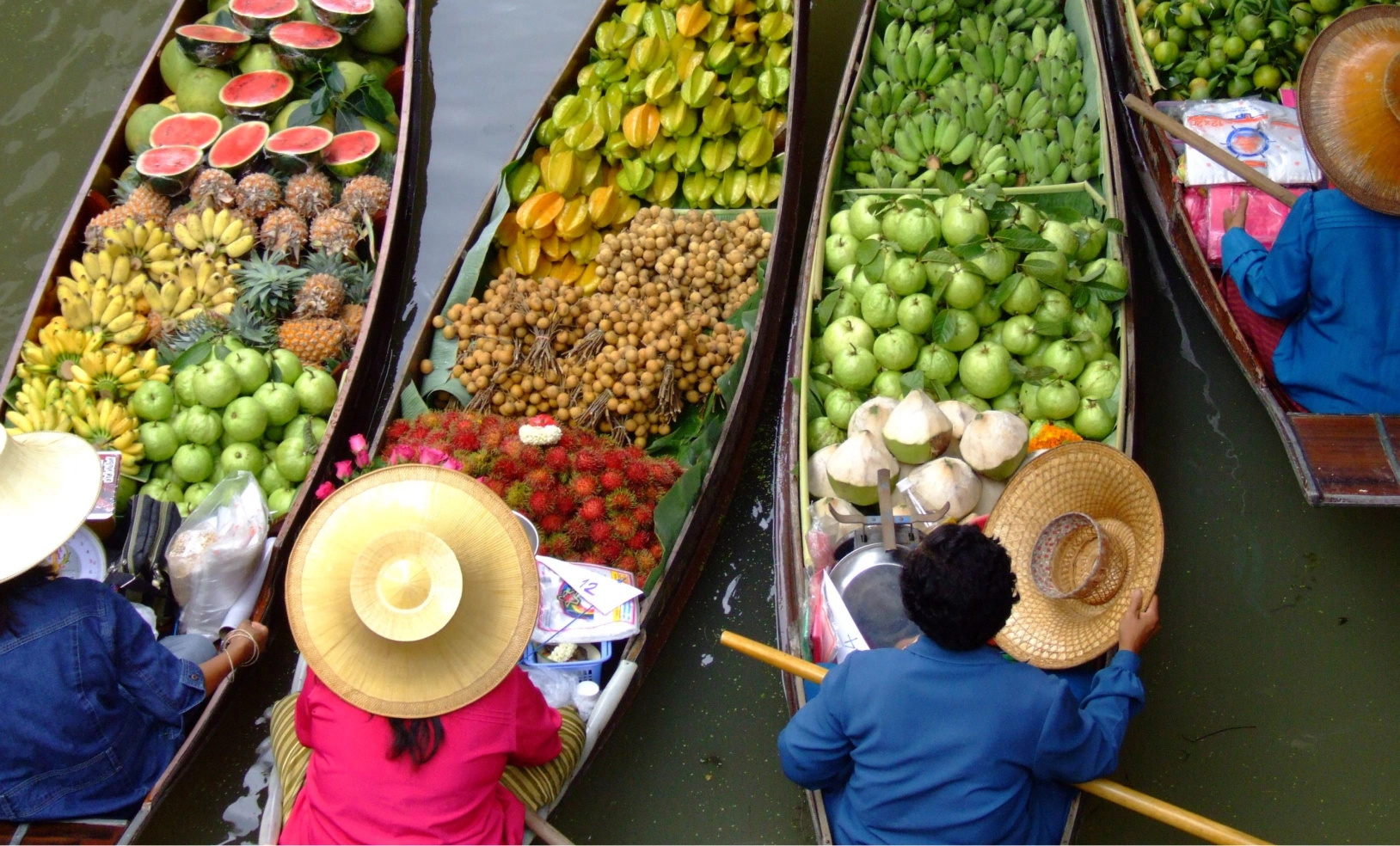 A high-angle view of several small wooden boats in a canal, each filled with colorful fresh produce managed by vendors in traditional hats.