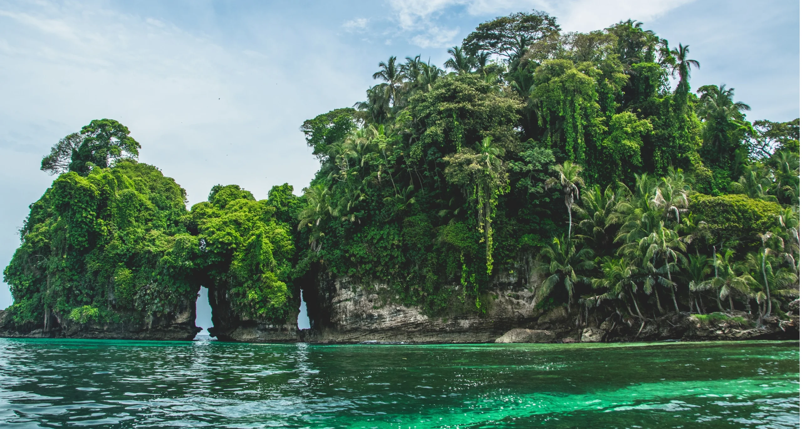 A rocky, tree-covered island with a small natural archway sitting in vibrant turquoise water under a bright sky.