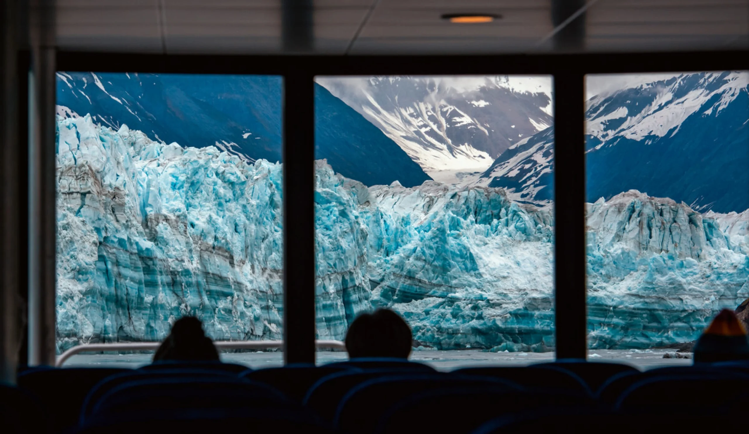 Two passengers looking out of a massive floor-to-ceiling window at a massive blue glacier.