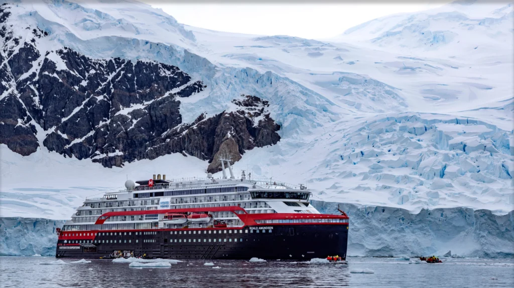 A sleek red and white HX expedition ship navigating near an ice-covered shore.