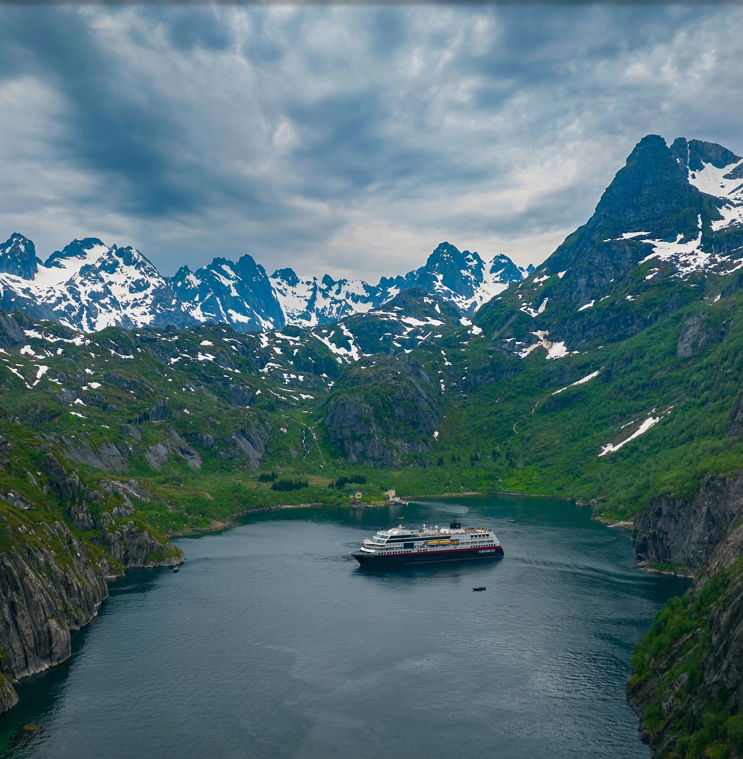 Aerial view of a ship navigating a narrow, turquoise fjord surrounded by high peaks.