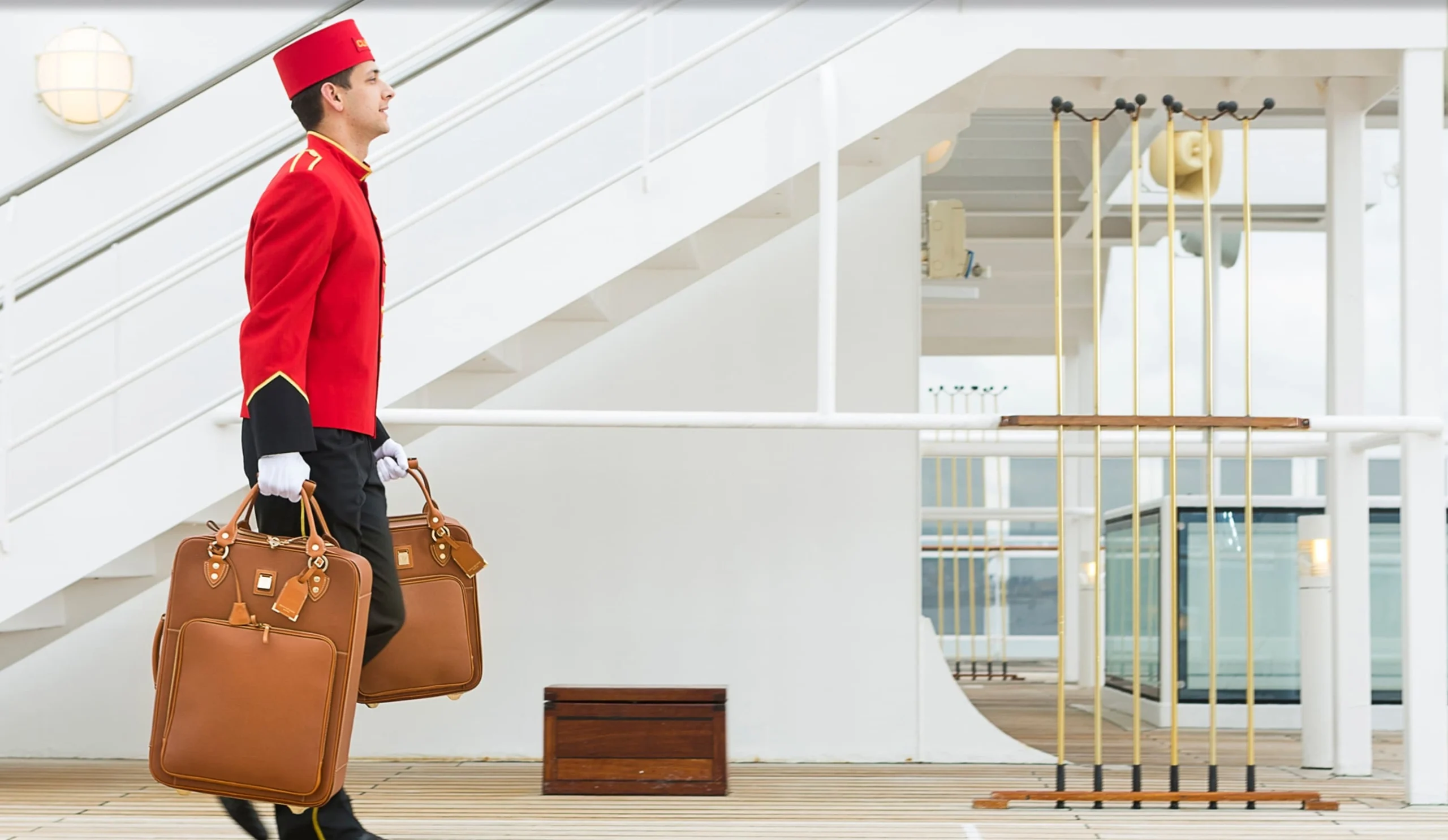 A porter in a crisp white and red uniform carrying leather luggage up a ship's gangway.