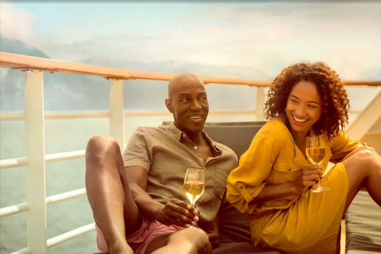 A couple laughing and holding glasses of white wine on a sunny ship balcony.