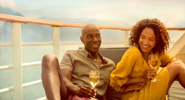 A couple laughing and holding glasses of white wine on a sunny ship balcony.