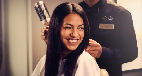 A woman smiling while getting her hair styled by a professional onboard stylist.