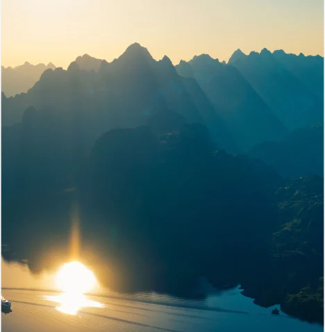 Hurtigruten Norwegian Fjords Cruises | Cruise Professionals Sunrise over Norway's majestic fjords viewed from a Hurtigruten cruise, surrounded by dramatic mountains and tranquil waters.