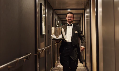 A ship crew member in a service uniform standing in a well-appointed stateroom doorway.
