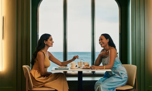 Two women laughing and talking while dining at a small table next to a large window.